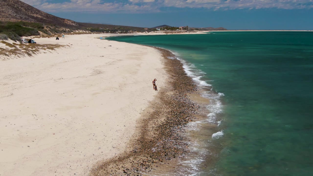 At Barakas Beach in Baja California Sur, a distant person walks along the rocky line where white sand meets turquoise water near the start of the clip.