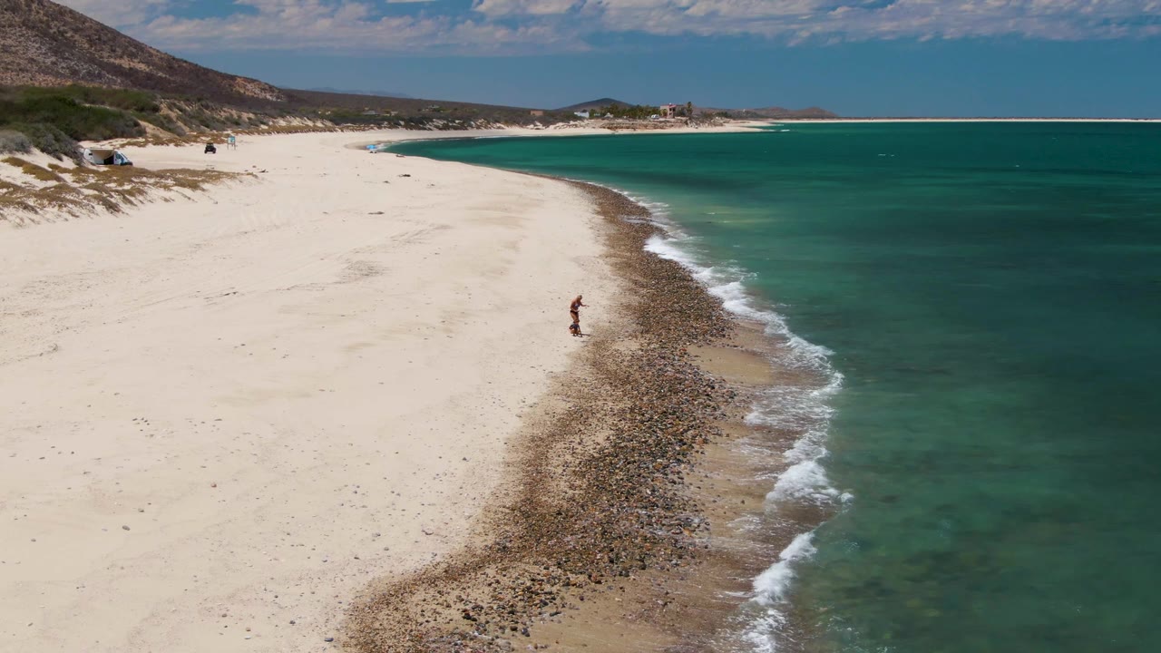 High aerial beach view with a lone person near the rocky shoreline and clear turquoise water.