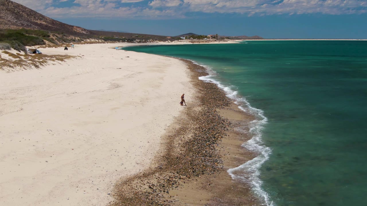The person appears slightly farther along the shoreline as small white waves break against the rocky strip beside the turquoise ocean at Barakas Beach.