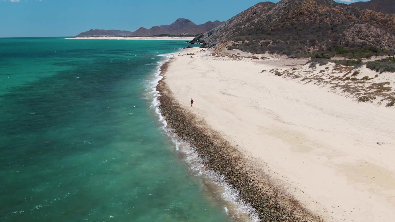 Aerial view at Barakas Beach in Baja California Sur with turquoise water on the left, white sand curving along the shore, rocky hills to the right, and a small person visible near the surf line.