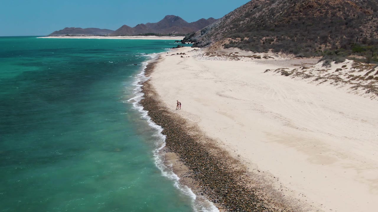 Drone view at Barakas Beach with two small people on the pale sand near a dark shoreline band, clear turquoise water to the left, and rocky Baja California Sur hills filling the right side.