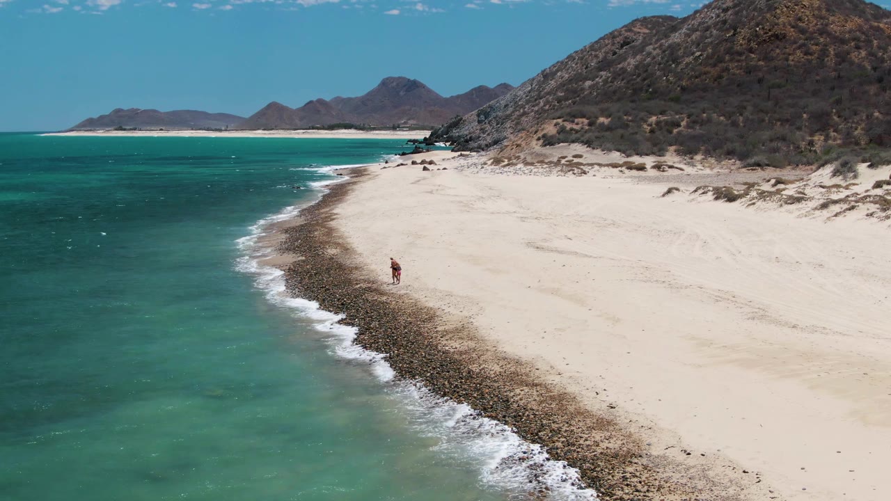 Aerial frame at Barakas Beach, Baja California Sur, showing turquoise ocean on the left, a curved rocky surf line, pale sand, mountains, and a small person near the shoreline.