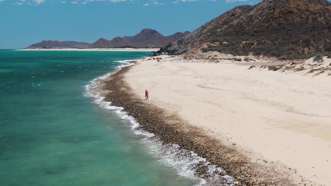 Drone view of a small person walking along a rocky edge of a sandy beach.