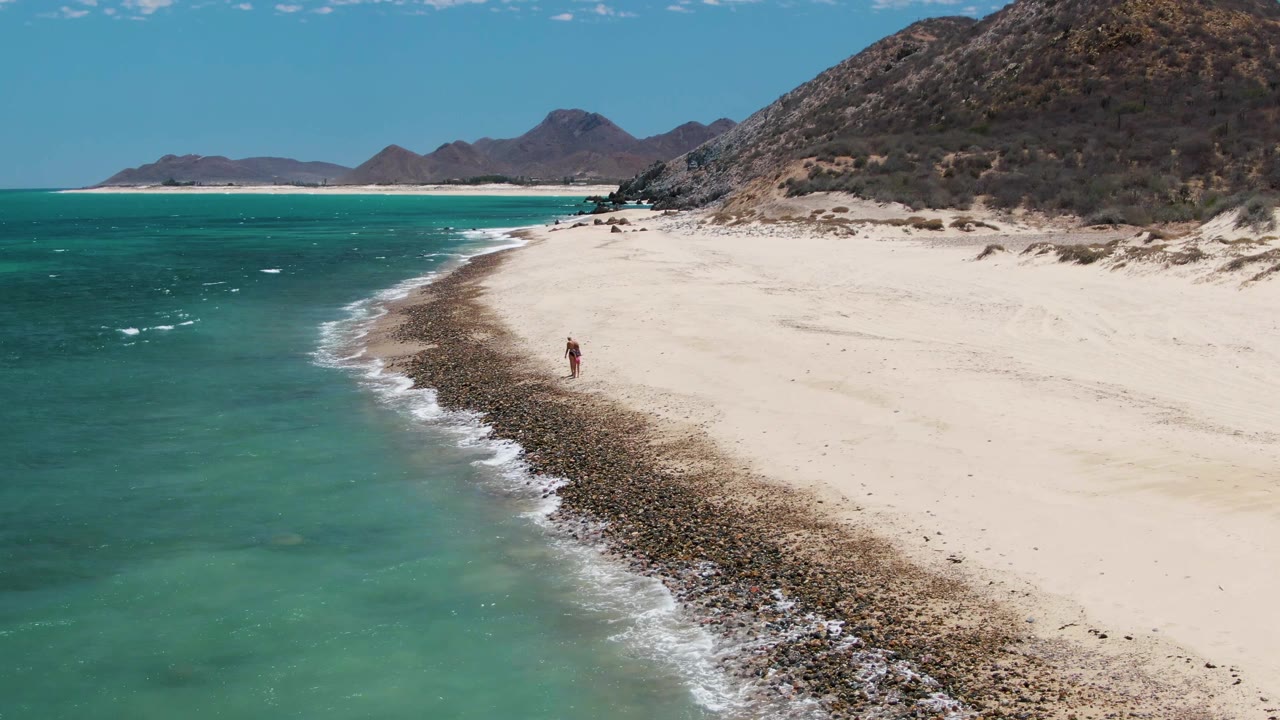 At 0:02, Barakas Beach appears from above with the small walker near center-right, a foamy wave line along the rocks, pale sand to the right, and mountains beyond the shore.