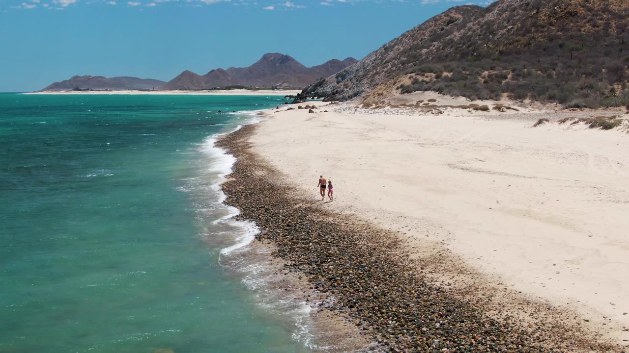 At Barakas Beach in Baja California Sur, two people walk along the curve where turquoise water meets pale sand and a dark rocky tide line.
