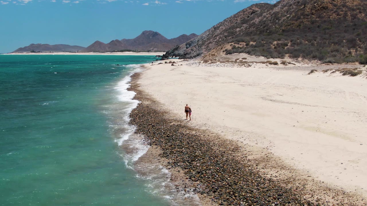 One person stands near the rocky shoreline as turquoise waves reach the sand.