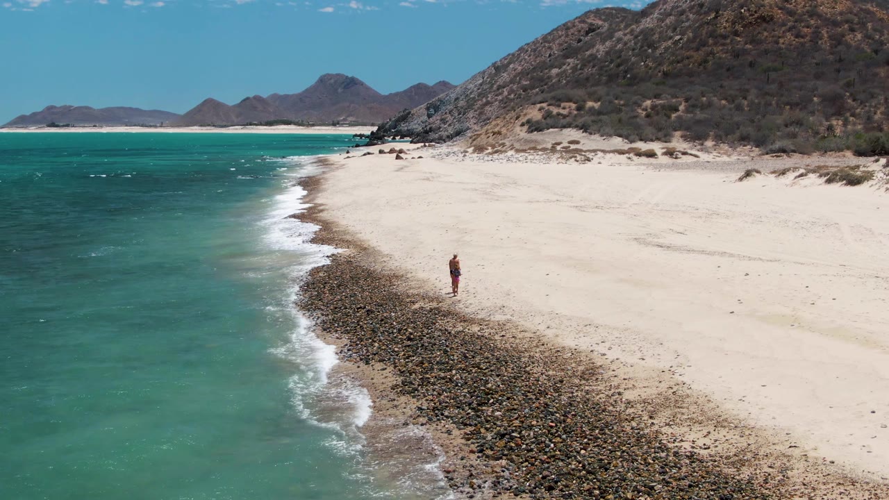 At Barakas Beach in Baja California Sur, a lone walker appears near the center-right of the beach beside the curving rocky tide line and turquoise surf.