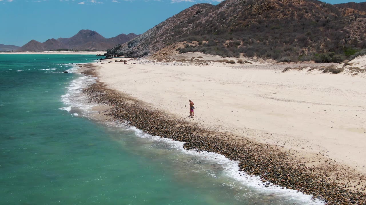 At Barakas Beach in Baja California Sur, one person walks near the waterline as turquoise ocean meets a rocky strip on the pale sand.