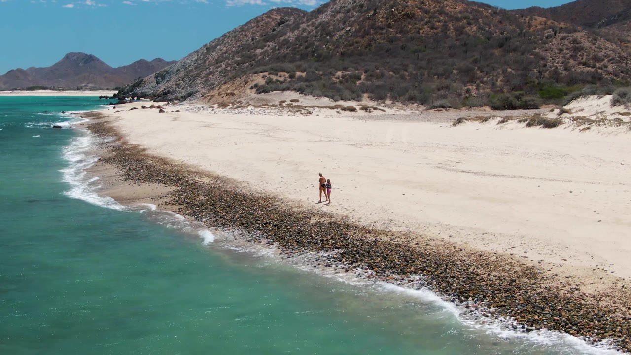 Two people are visible on the sand at Barakas Beach, walking beside the curved rocky strip with turquoise water on the left and hills behind.