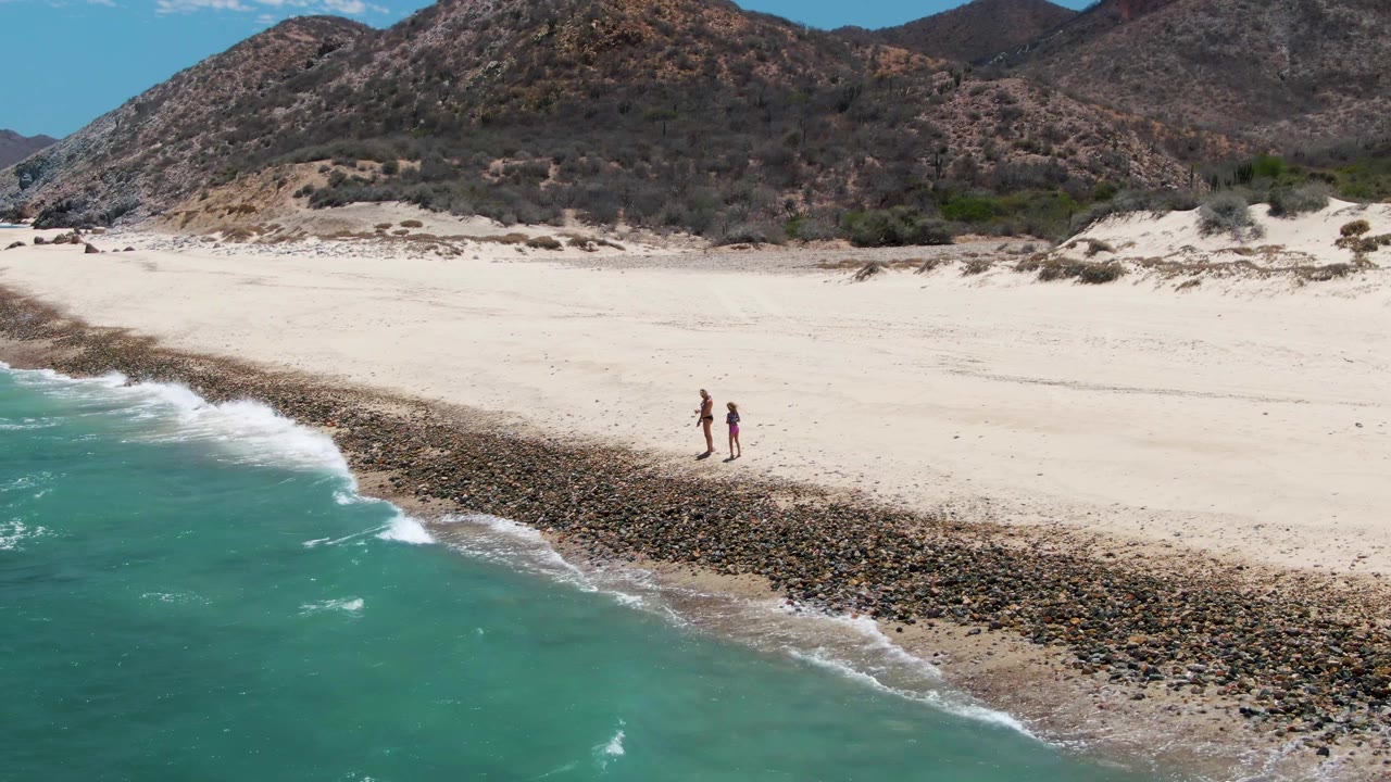 At Barakas Beach in Baja California Sur, two people walk along pale sand near a rocky tide line as turquoise water fills the lower left of the frame.