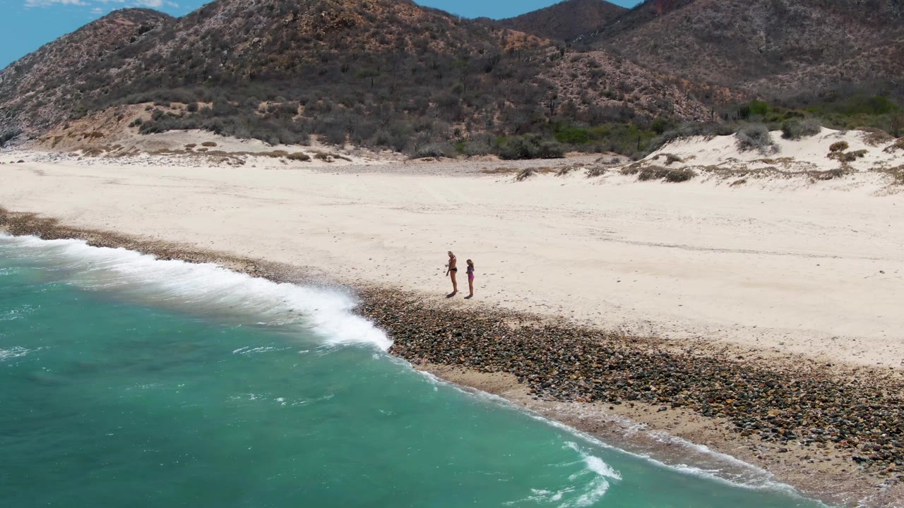 Two walkers on open sand with waves breaking along a dark rocky shore.
