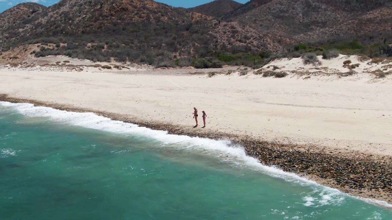 At Barakas Beach in Baja California Sur, two people appear near the center-right of the wide beach as foamy turquoise surf pushes farther up the rocky shoreline.