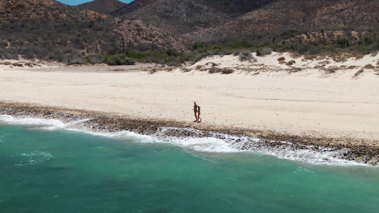 Two people walk near the shoreline at Barakas Beach in Baja California Sur, Mexico, with turquoise water in the foreground and mountains behind the pale sand.