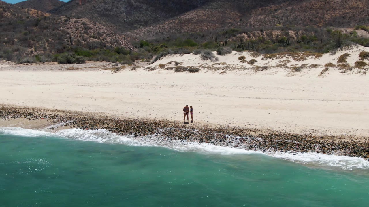 Two small figures remain near the center of the Barakas Beach shoreline, framed by clear turquoise ocean below and scrub-covered dunes before the mountains above.