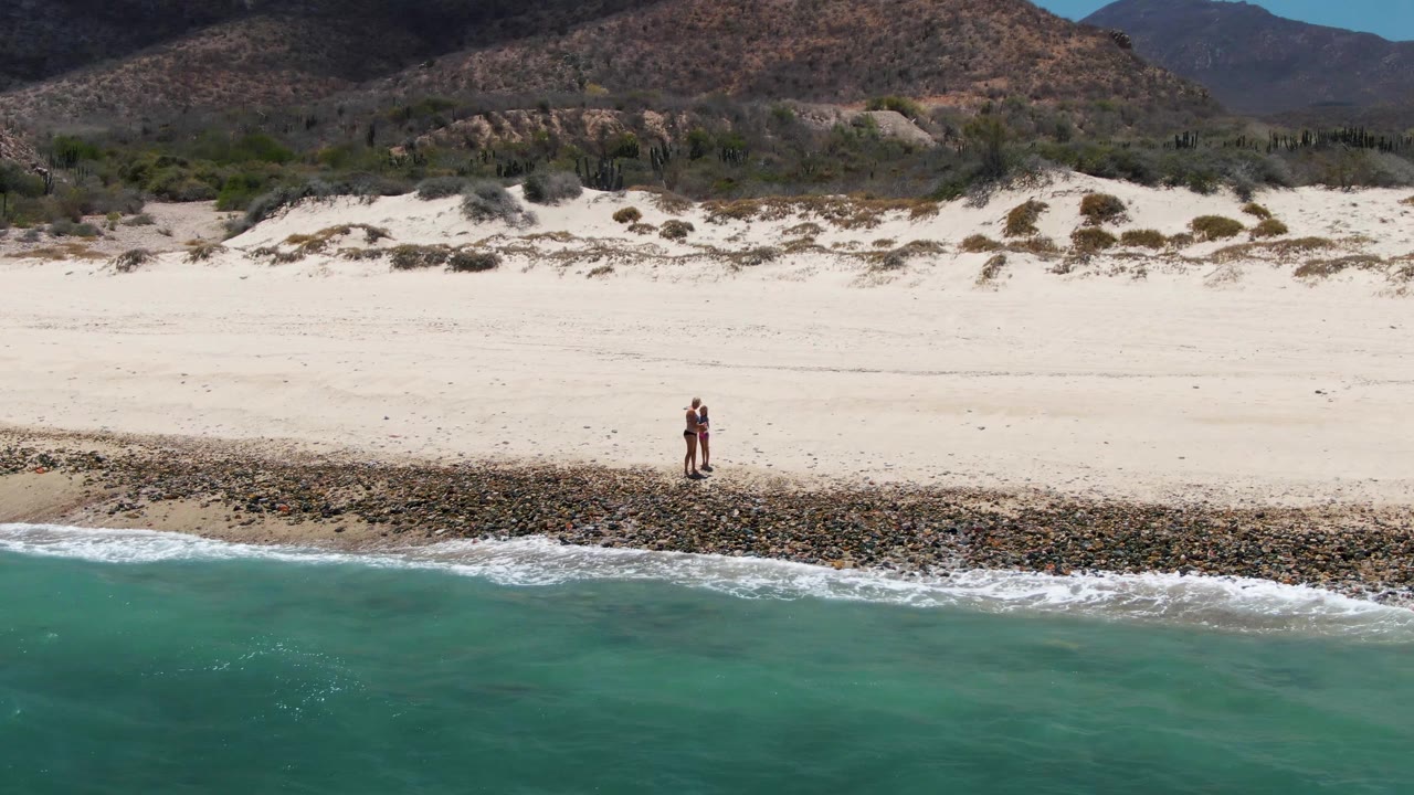 At Barakas Beach in Baja California Sur, a lone person stands or walks near the rocky shoreline as turquoise water fills the foreground and dunes rise toward mountains behind.