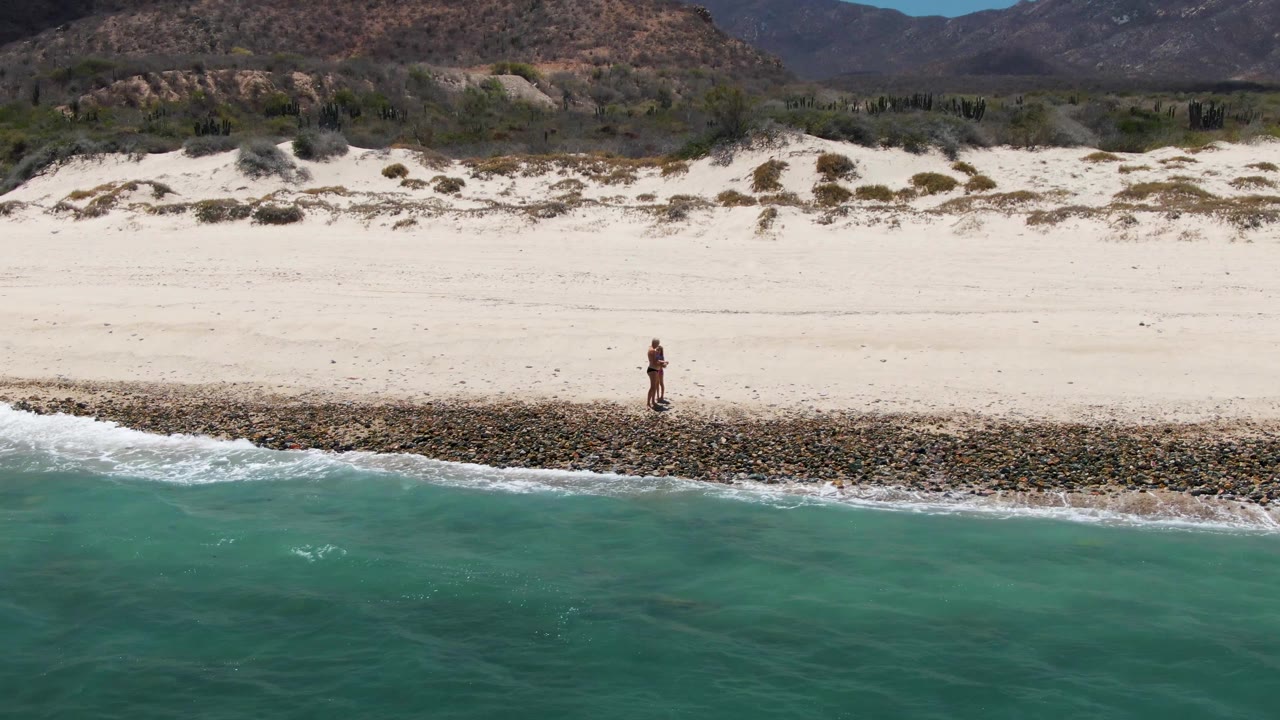 At 0:02 on Barakas Beach, the lone walker appears near the right side of the shoreline while turquoise water and a fresh wave line push toward the rocks.