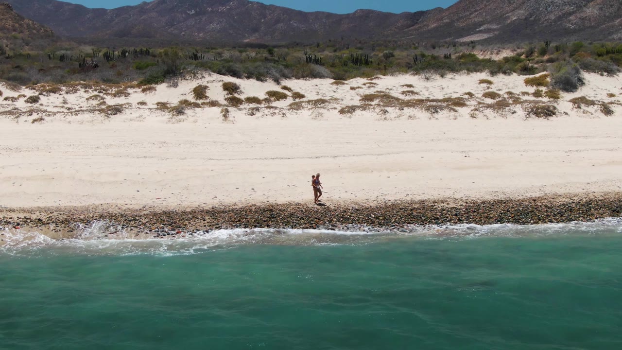 Two people walk along the rocky edge of Barakas Beach in Baja California Sur, with turquoise water below the frame and pale dunes and mountains behind them.