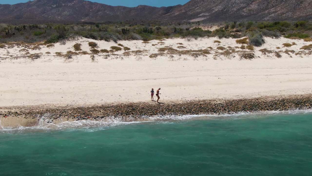 Two walkers on pale sand near a rocky shoreline and clear turquoise water.