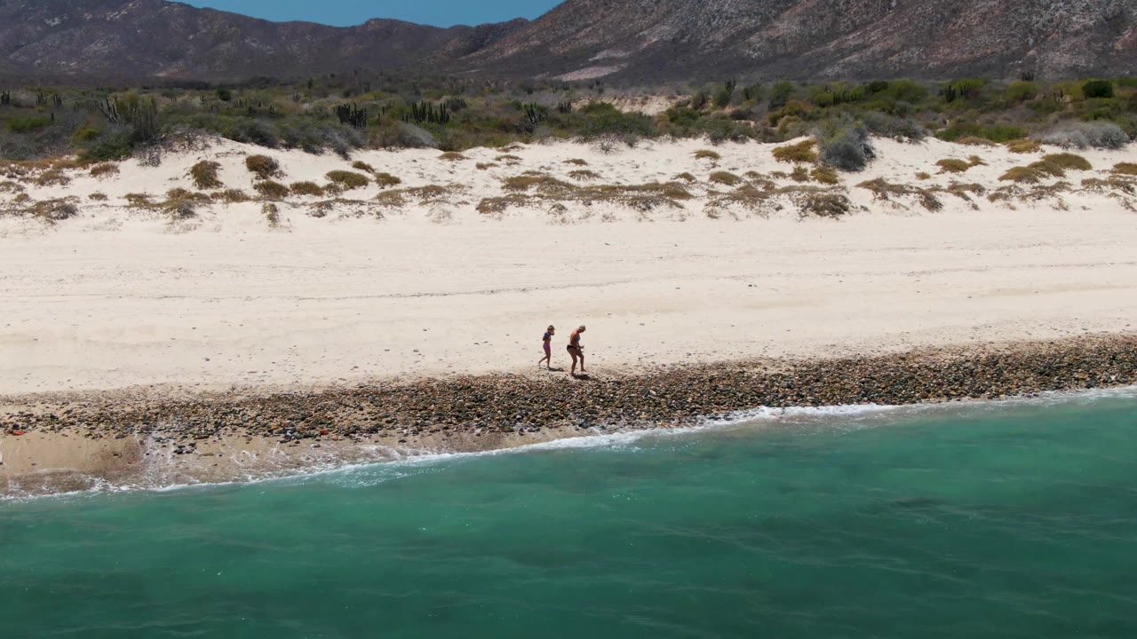 Two small figures walk beside the rocky surf line at Barakas Beach, with bright sand above them, green-blue water below, and dry mountains across the horizon.