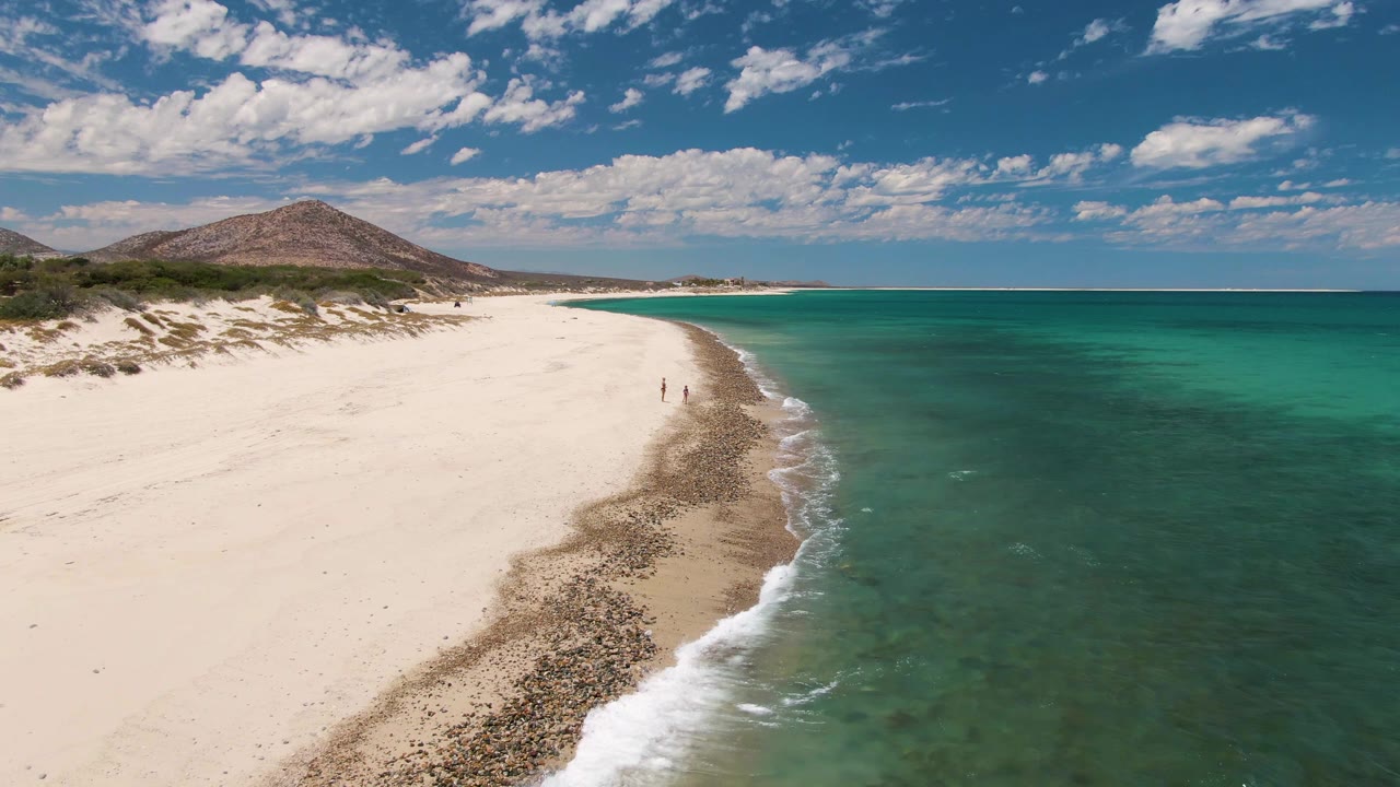 Opening aerial frame of Barakas Beach, Baja California Sur, with white sand on the left, turquoise ocean on the right, a rounded hill inland, and two tiny people near the shoreline.