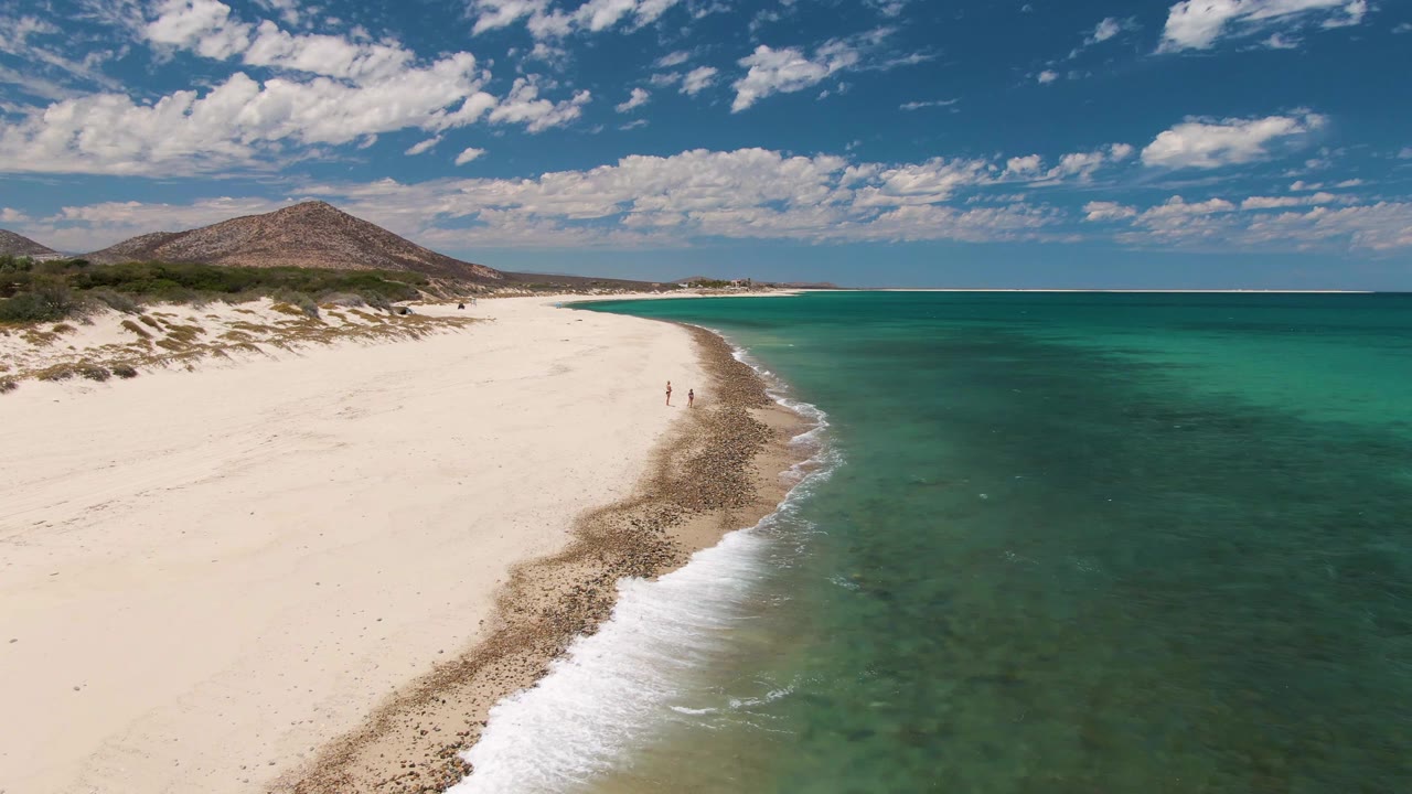 At 0:01, Barakas Beach appears from above with a brighter strip of foam advancing along the curved shore, two small figures near the water, and open turquoise water filling the right side.