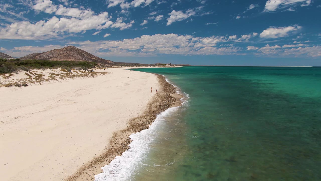 At 0:02, the aerial view holds on Barakas Beach as the foam thins along the waterline, two small people stand near mid-beach, and the inland hill and cloud bands remain in the background.