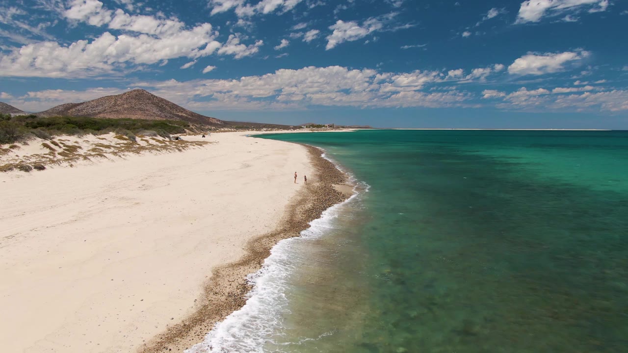 Wide aerial view of sandy beach, green-blue water, and inland hills.