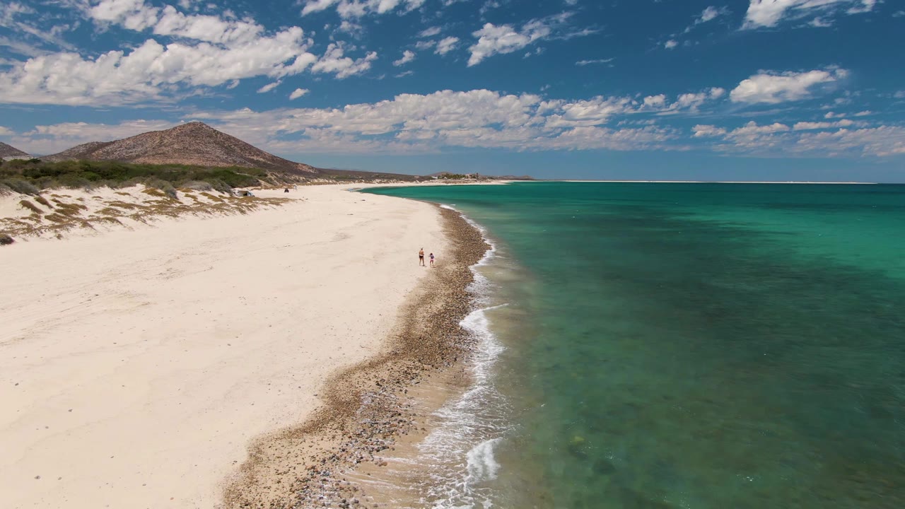 Aerial view of Barakas Beach with white sand on the left, turquoise ocean on the right, a curved wet shoreline, and two small people walking near the surf at the start of the clip.