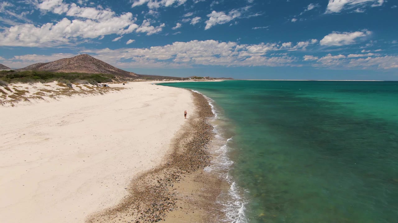 Aerial frame of Barakas Beach with one small visible person near the shoreline, clear green-blue water, pale sand, low dune vegetation, and a rounded hill under scattered clouds.