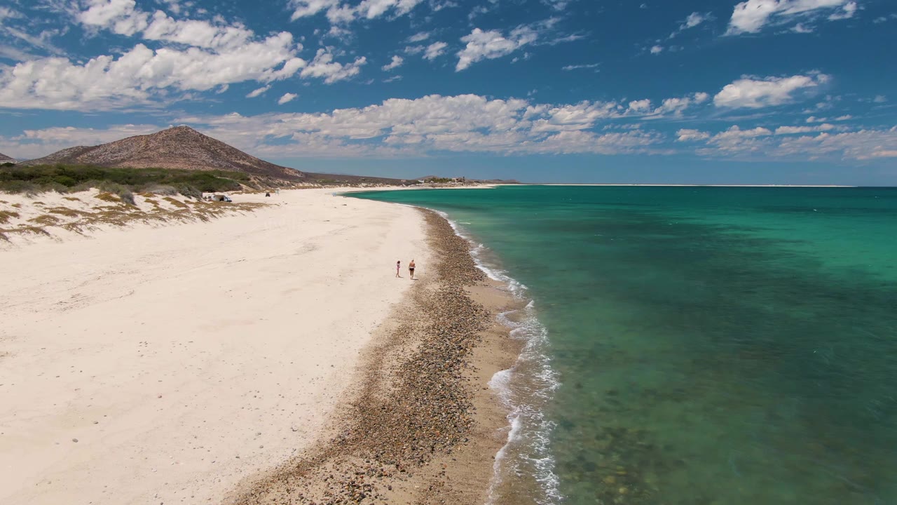 Aerial view at Barakas Beach, Baja California Sur, showing two people walking near the waterline as white sand curves toward a low hill under scattered clouds.