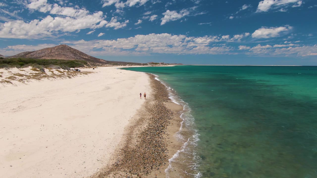 Two small figures walk along the beach edge with turquoise ocean to the right.