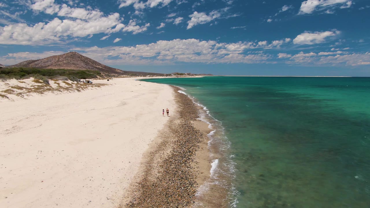 At 0:02, two walkers remain visible near the center of the Barakas Beach sand, with clear green-blue water on the right and a low hill beyond the dunes.