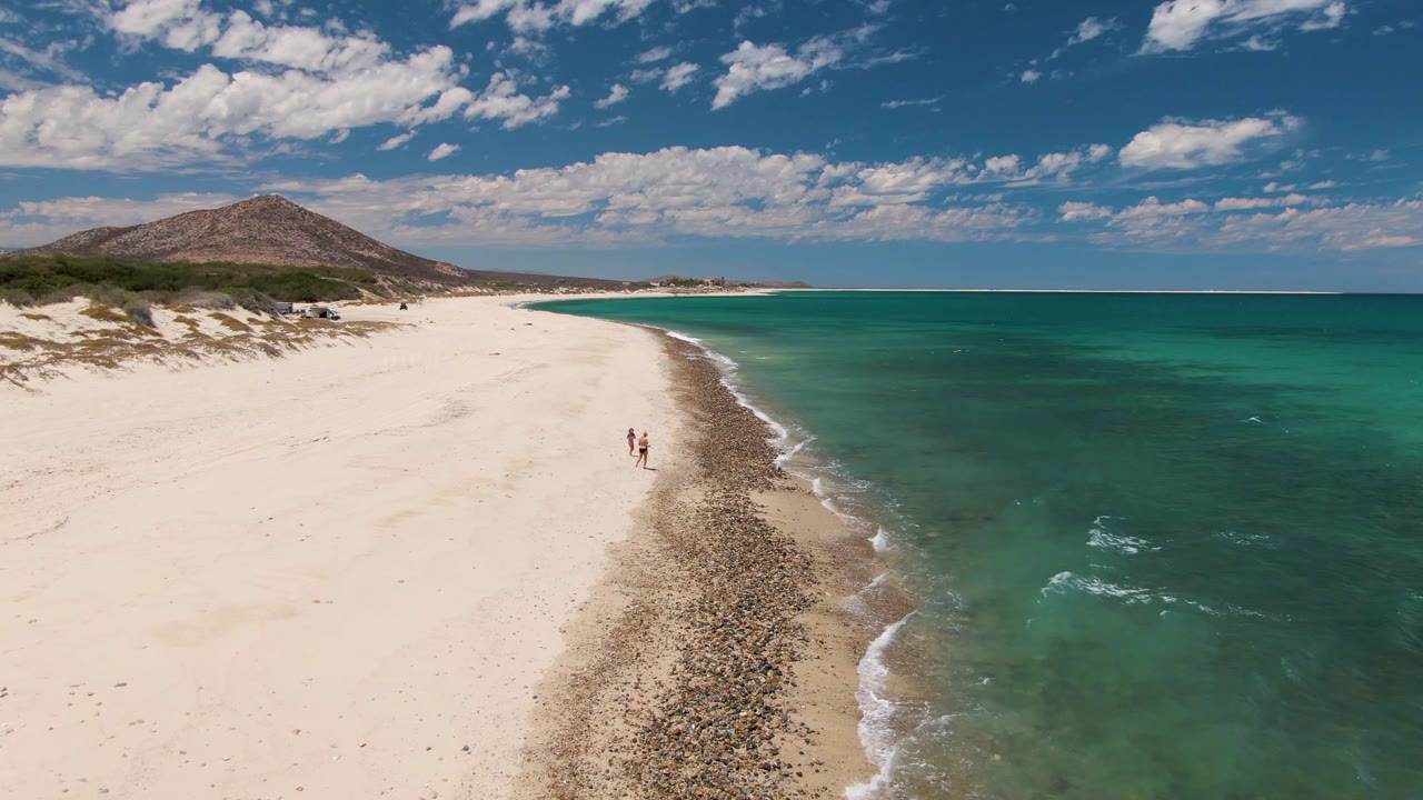 Aerial frame at Barakas Beach, Baja California Sur, showing two people walking along a white sand shoreline with turquoise ocean to the right and a low hill beyond the beach.
