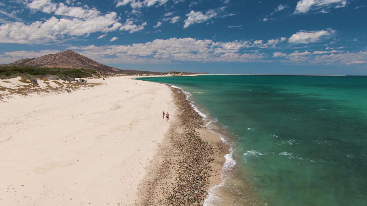 Two walkers on a pale beach with a curved rocky waterline and turquoise sea.