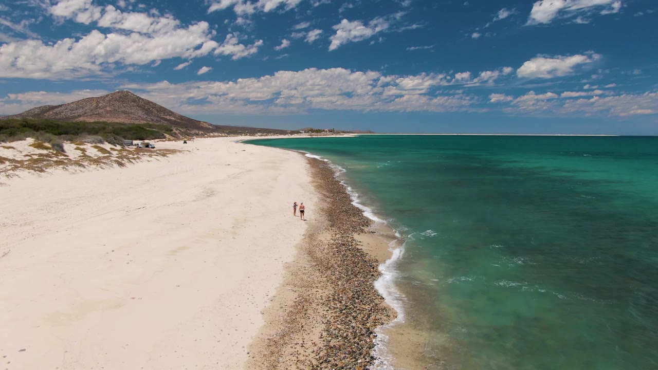 At Barakas Beach, two people remain visible beside the foamy edge of the turquoise ocean, with a broad white sand beach, scattered beach vegetation, blue sky, and a low background hill.