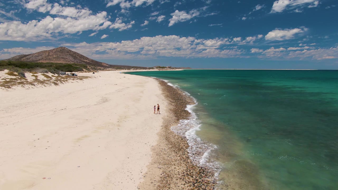 Aerial frame from Barakas Beach in Baja California Sur showing two people on pale sand near a darker shoreline band, with turquoise ocean on the right and a low hill in the distance.