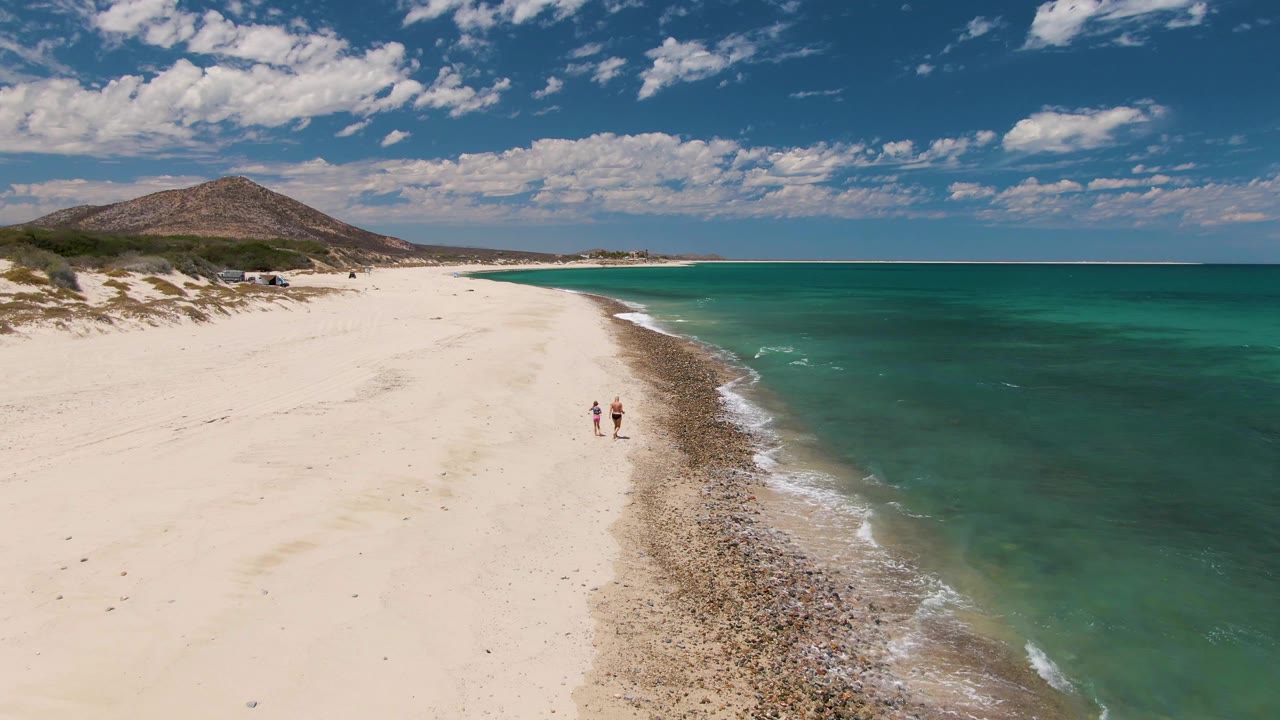 At 0:02, two people are visible on Barakas Beach near the curved shoreline, while clear shallow water and a thin band of white wave foam mark the right edge of the sand.