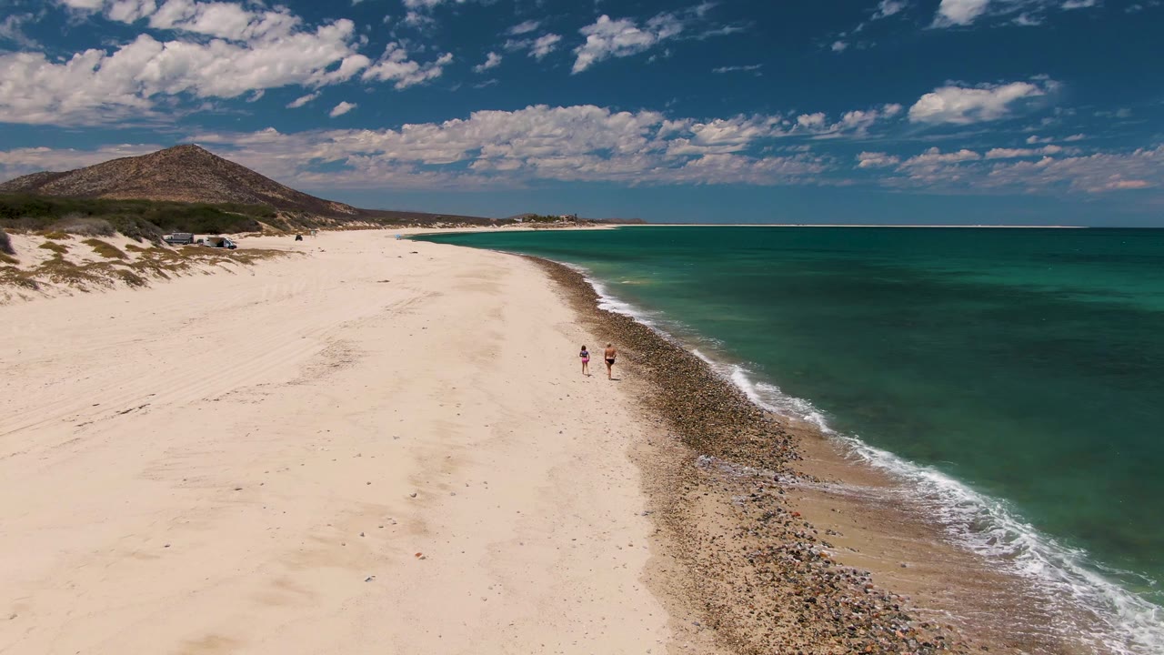 Two people walk along the sandy shoreline with ocean water beside them.
