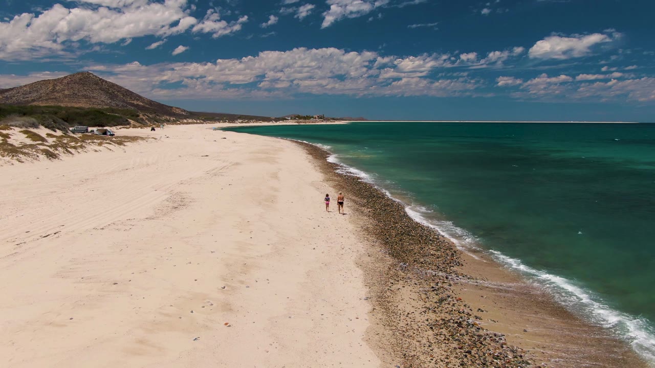 Aerial frame from Barakas Beach showing two walkers slightly farther along the curving shoreline, with the turquoise sea taking the right half of the image.