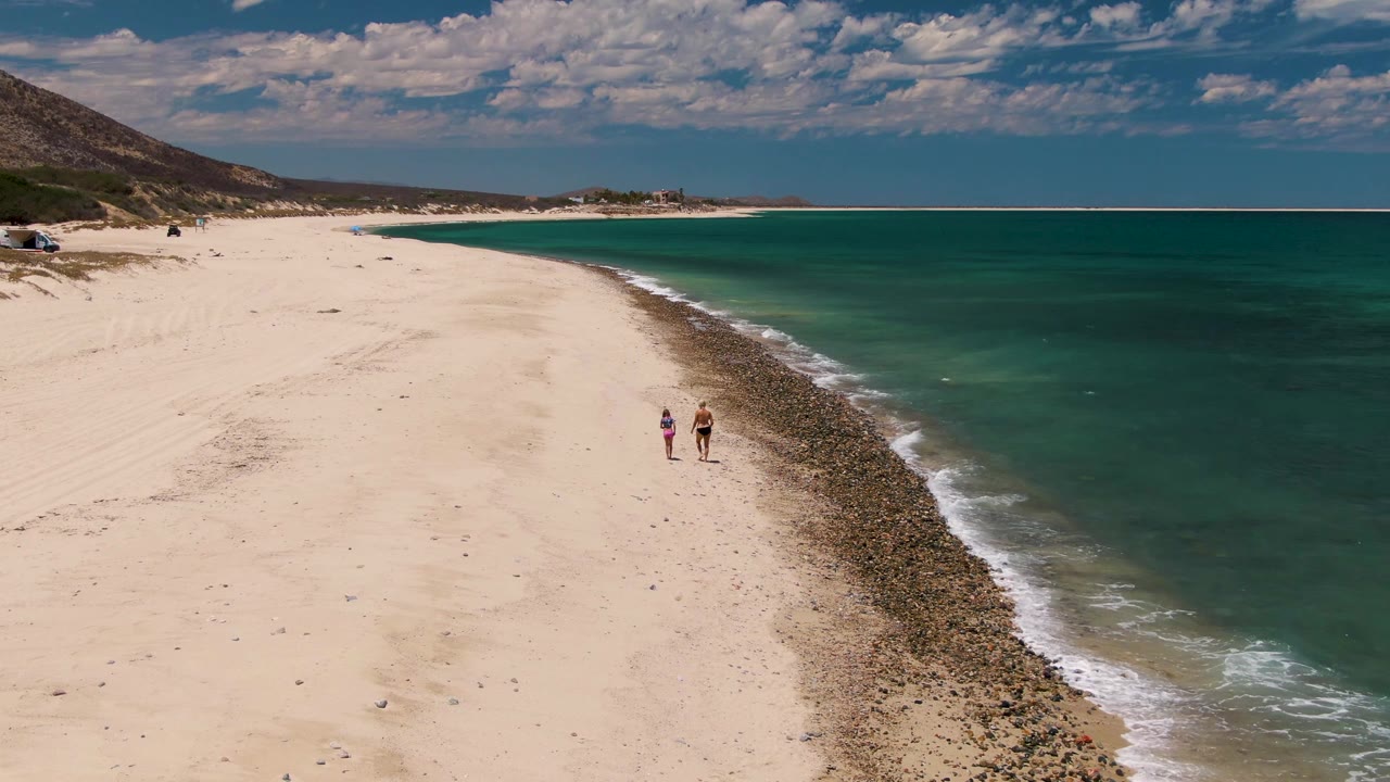 Two people walk along Barakas Beach in Baja California Sur, with pale sand on the left, turquoise ocean on the right, and a low hill under scattered clouds.