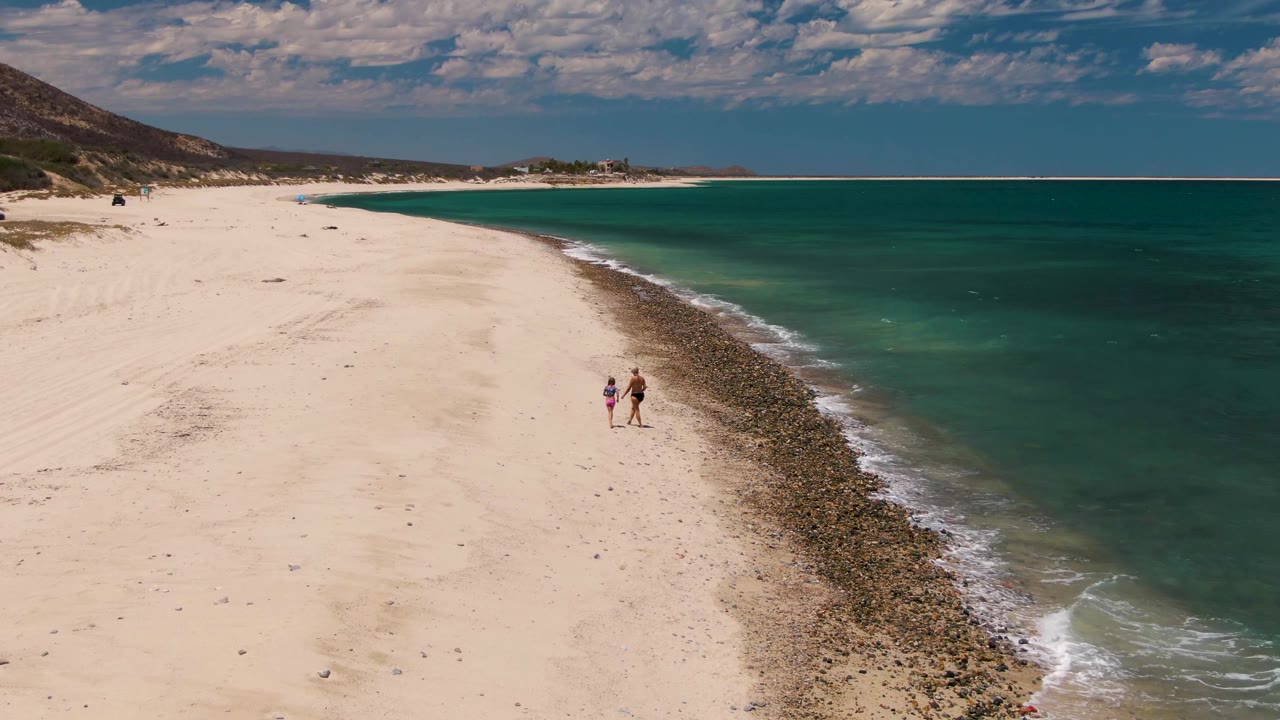 Aerial view of two walkers near waves on a pale sandy shore.
