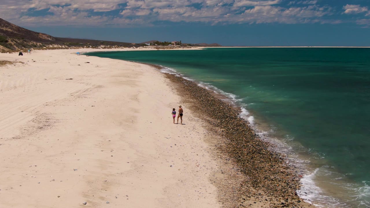 Two people appear slightly farther along the Barakas Beach shoreline, with the rocky strip bending through the frame and turquoise water filling the right side.