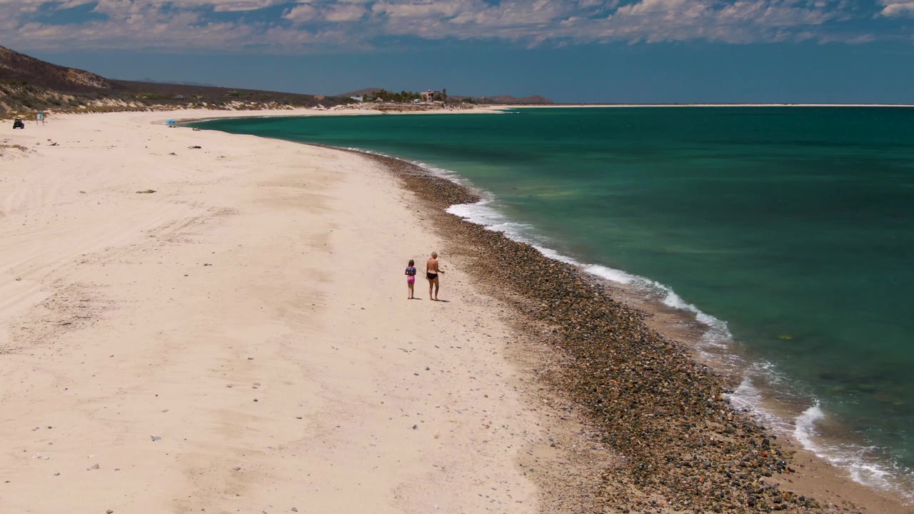 At Barakas Beach in Baja California Sur, two distant people walk along pale sand near a dark pebble band and turquoise ocean under scattered clouds.