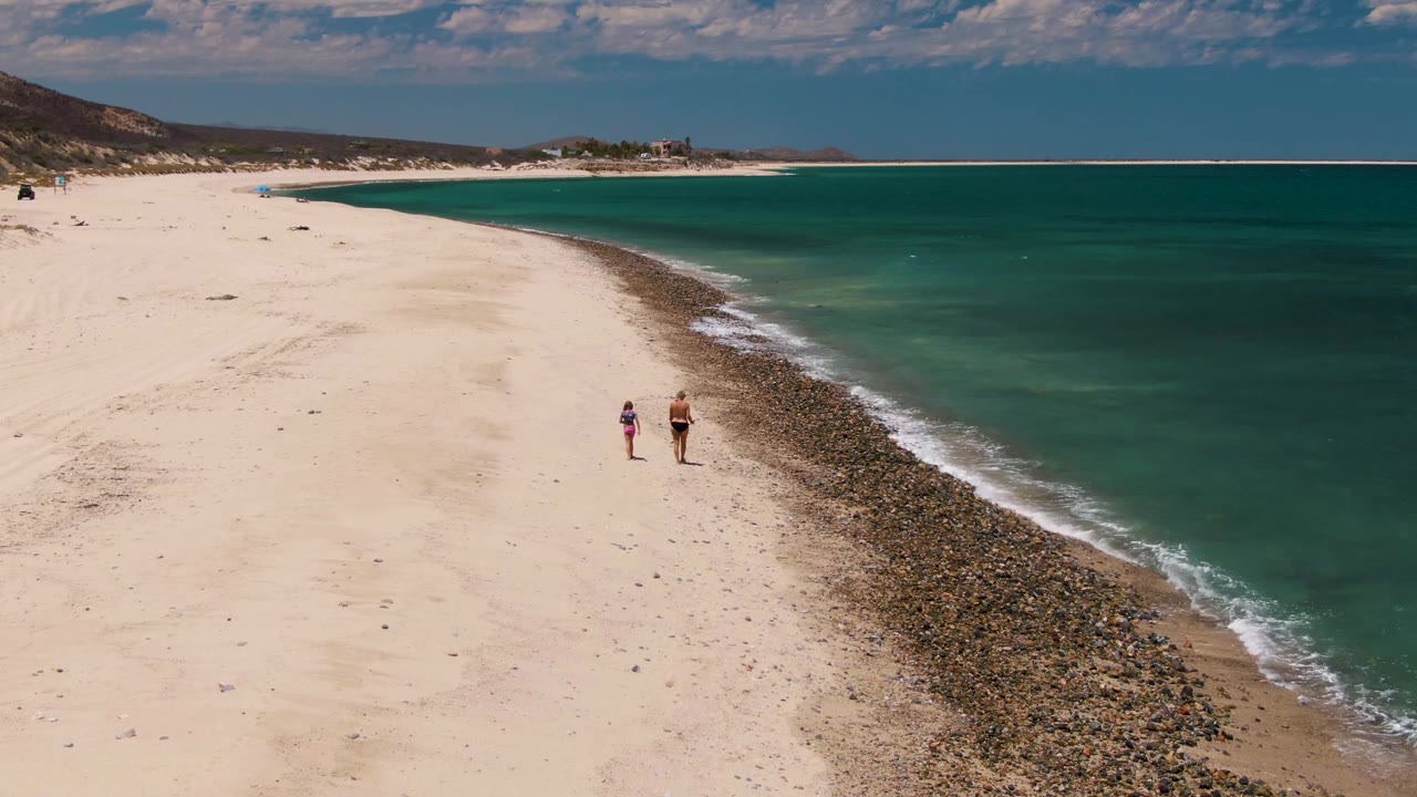 At 0:02, the two figures are slightly farther along the shoreline while the turquoise water deepens toward the right side of the Barakas Beach frame.