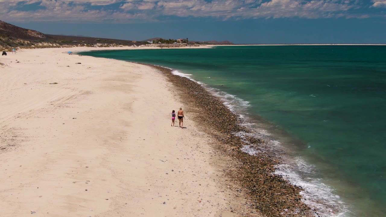 Two walkers near a rocky shoreline on a broad white sand beach.