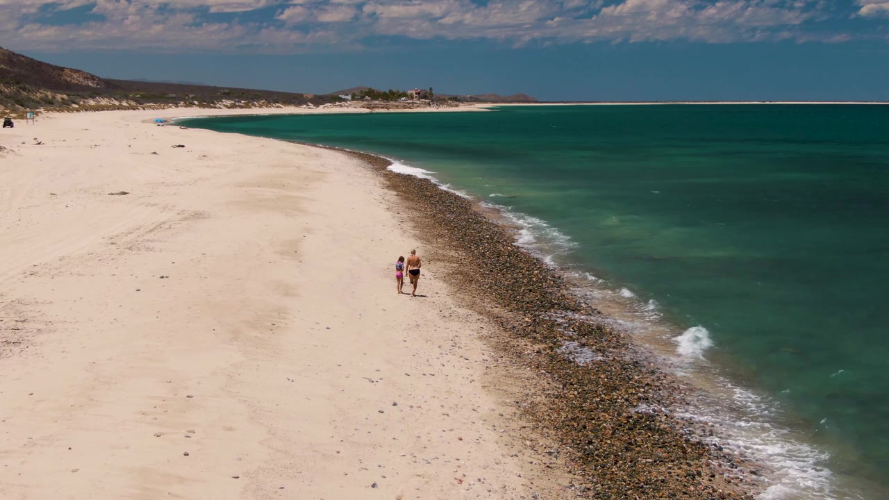 Aerial view at Barakas Beach with two people near the center-left, the rocky waterline below them, and a long turquoise bay stretching toward the horizon.