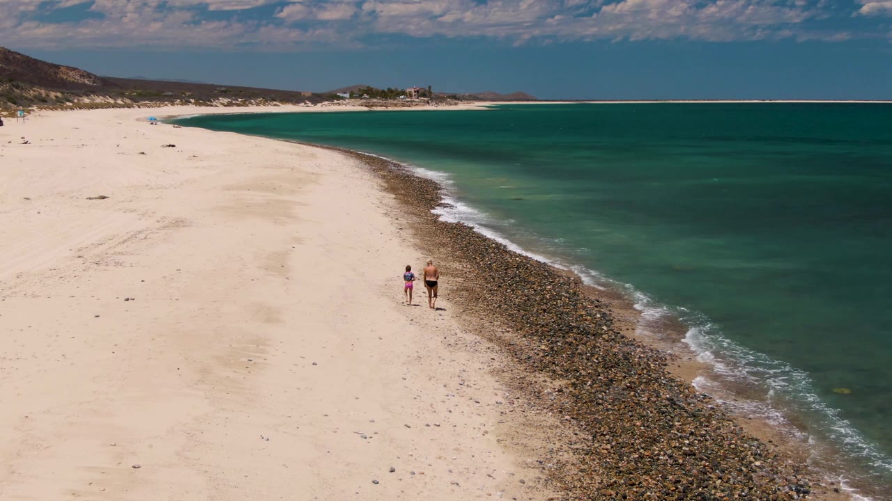Opening aerial frame over Barakas Beach in Baja California Sur, with two people on pale sand beside a dark pebble shoreline and turquoise ocean.
