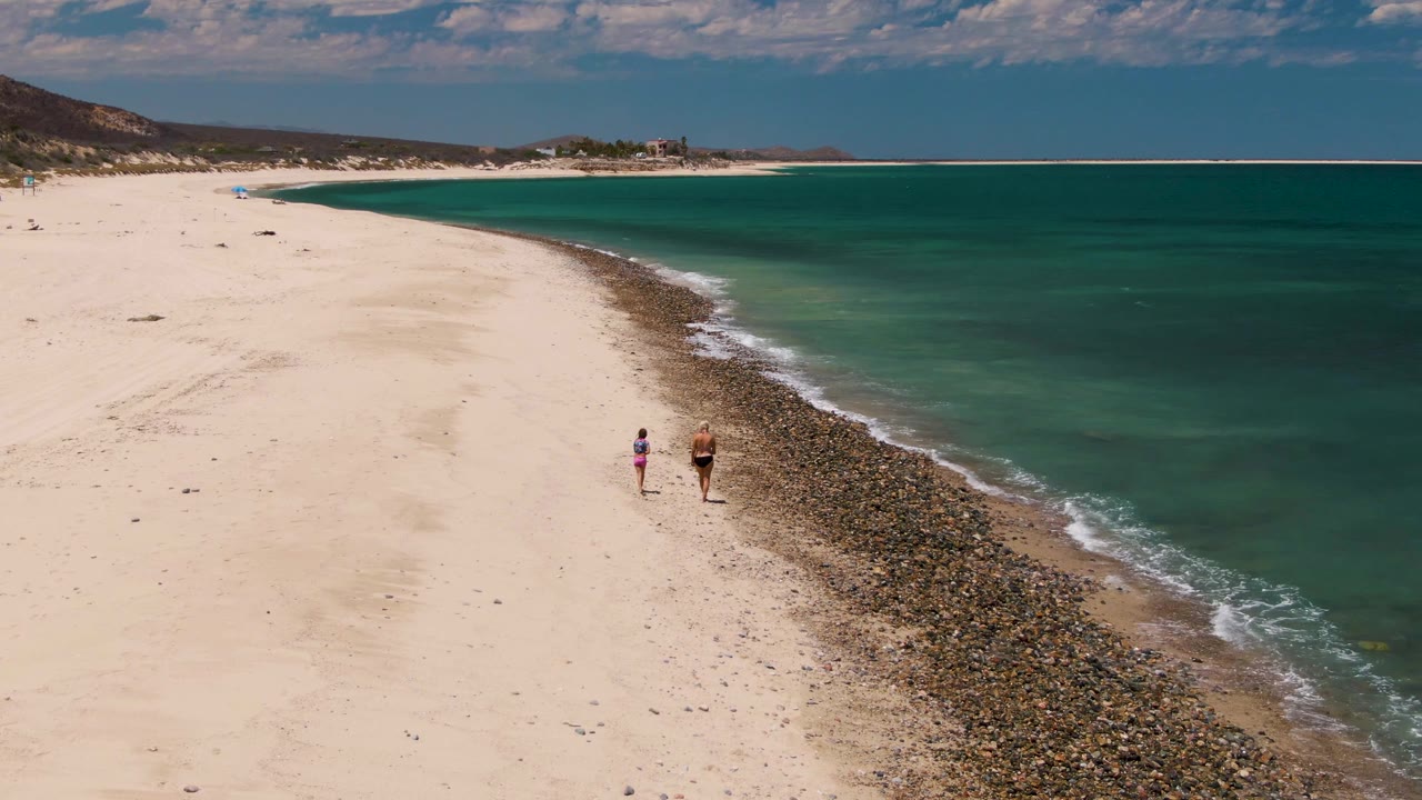 Two beachgoers walking along sand next to a pebbled surf line.