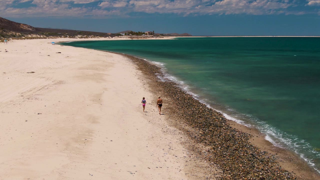 Mid-clip aerial frame at Barakas Beach with two small figures walking beside the curving dark shoreline and broad turquoise water filling the right side.