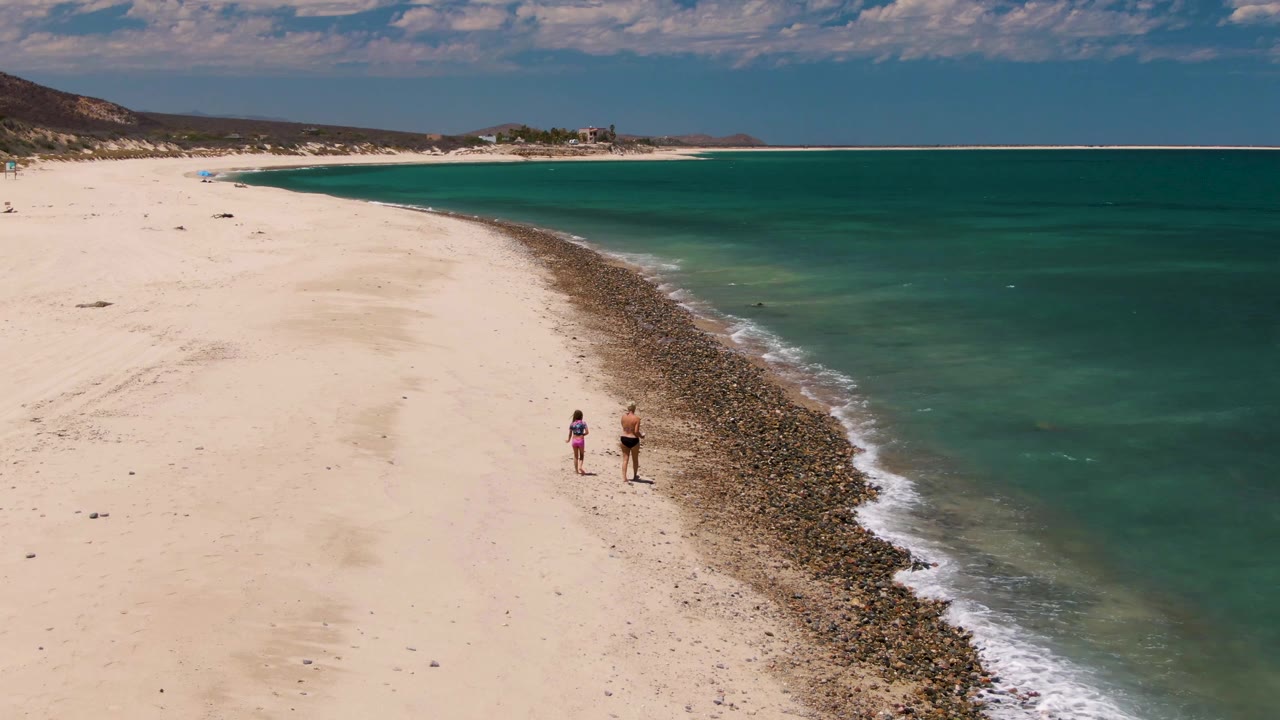 At Barakas Beach in Baja California Sur, two small figures walk along pale sand beside a diagonal band of rocks and turquoise water near the start of the clip.